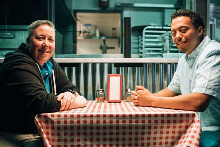 A photograph of two men sitting at a table in a diner.