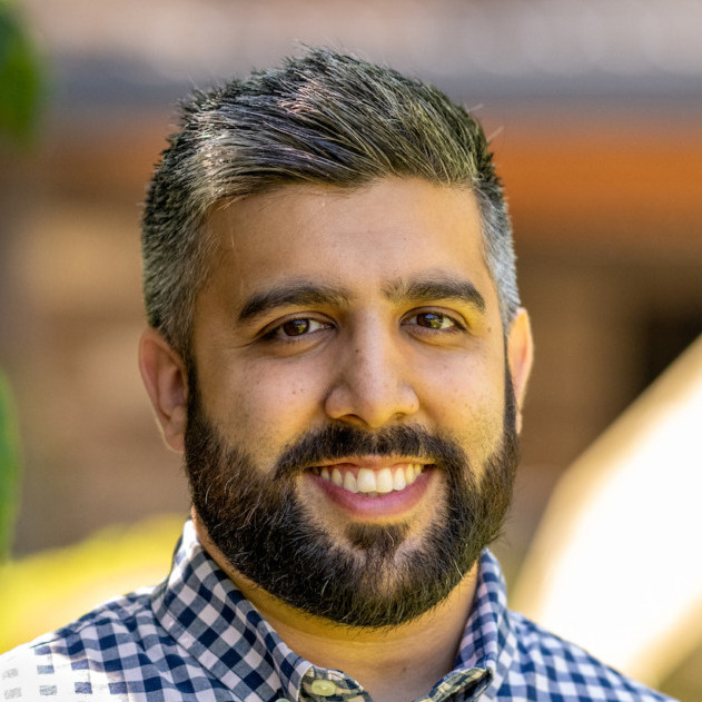 Zaki Barzinji's headshot. A person with short hair and a beard, wearing a collard shirt, is outside smiling at the camera.